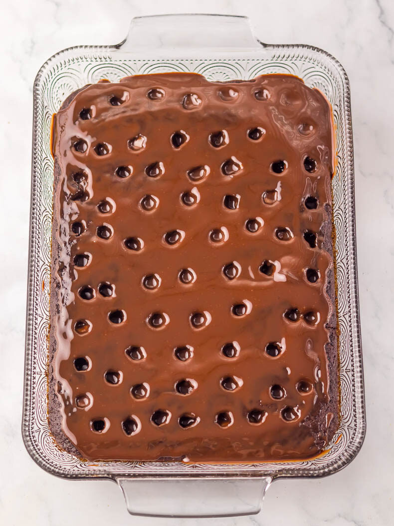 A rectangular dr pepper cake poke cake in a glass baking dish, with visible holes and topped with a smooth chocolate glaze.
