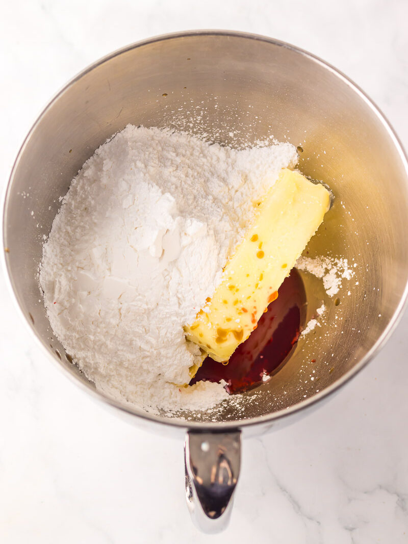 A mixing bowl containing powdered sugar, a stick of butter, and a splash of vanilla extract on a white surface.