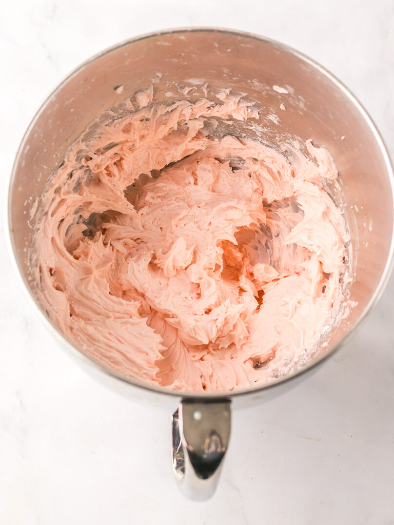 A mixing bowl filled with light pink whipped frosting on a white marble surface.