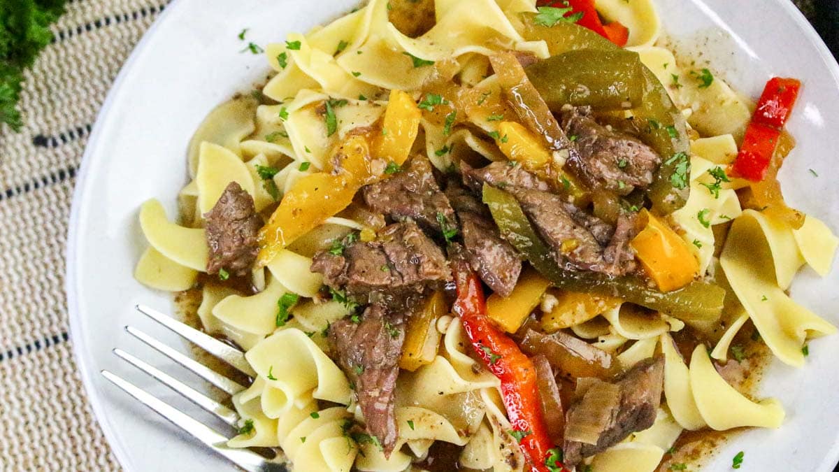 A plate of egg noodles topped with sliced beef, bell peppers, onions, and brown sauce, with a fork on the side.