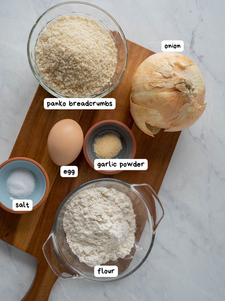 A wooden board with panko breadcrumbs, a whole onion, an egg, garlic powder, salt, and flour in separate bowls, each labeled.