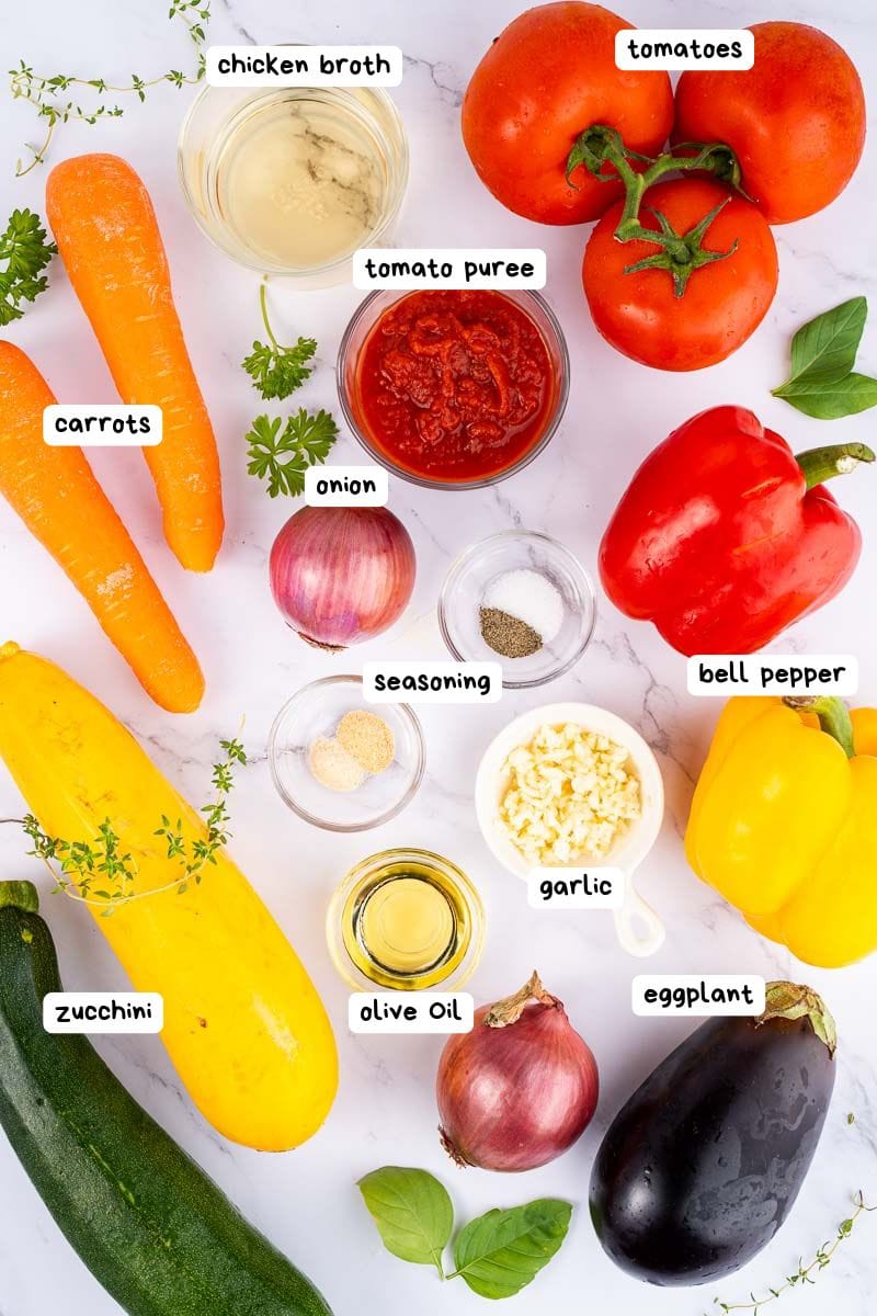 Assorted fresh vegetables, seasonings, olive oil, and chicken broth arranged on a white surface, each labeled with its name.