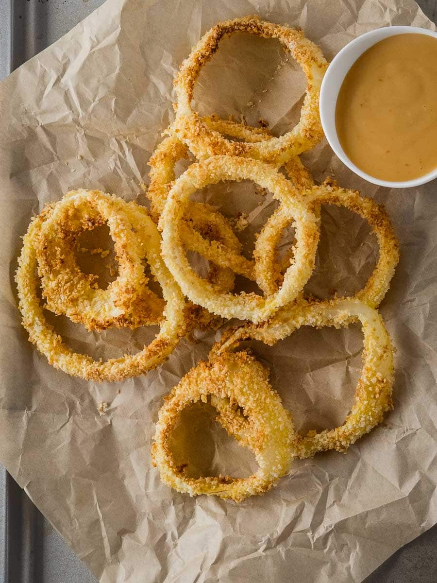 A pile of crispy breaded onion rings on brown parchment paper next to a small cup of dipping sauce.