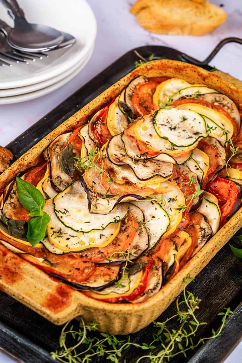 A baked ratatouille dish with thinly sliced zucchini, eggplant, squash, and tomato arranged in overlapping layers, garnished with fresh herbs.
