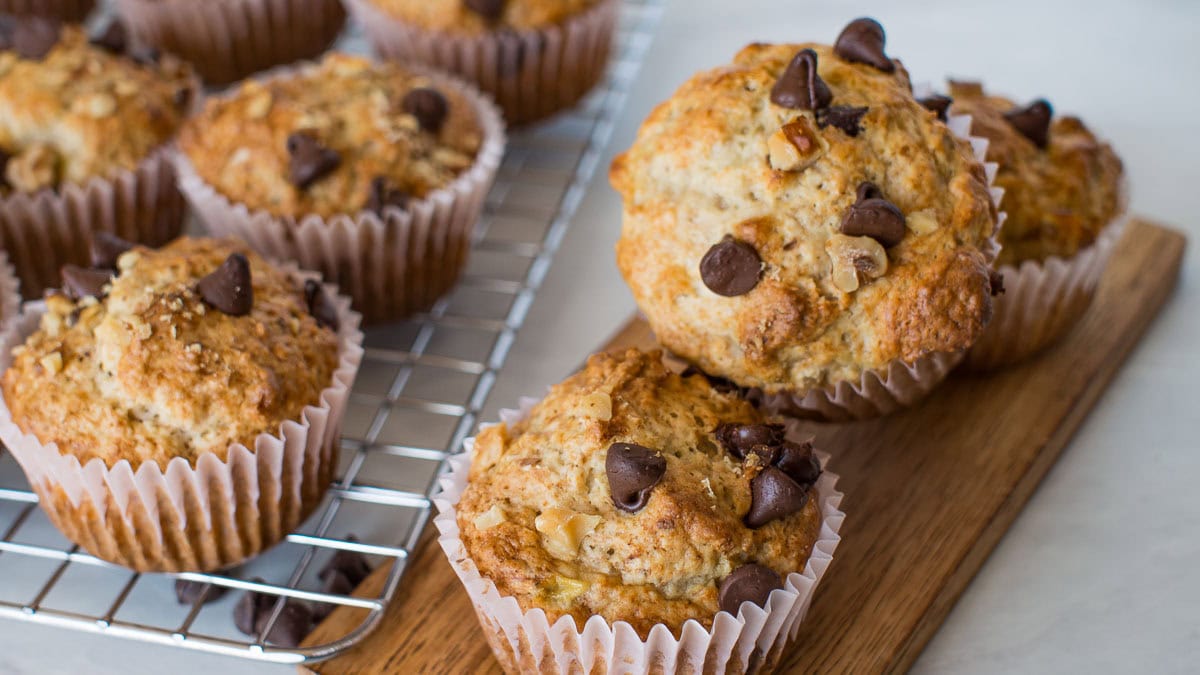 Chocolate chip muffins sit on a cooling rack and wooden board, with paper liners and a golden-brown top.