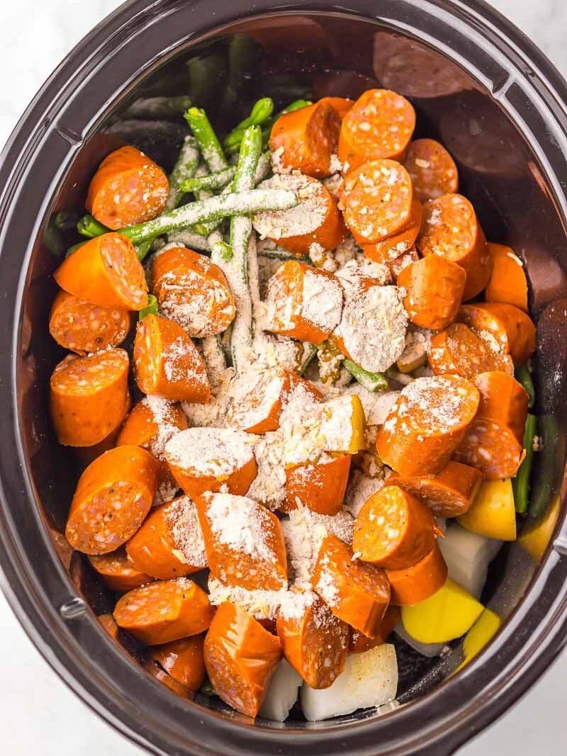 Sliced sausage, green beans, potatoes, and powdered seasoning in a slow cooker, ready to be cooked.
