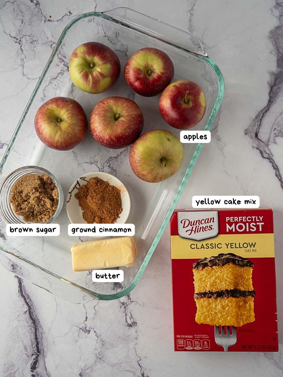 A glass baking dish with apples, brown sugar, ground cinnamon, butter, and a box of Duncan Hines classic yellow cake mix on a marble surface.