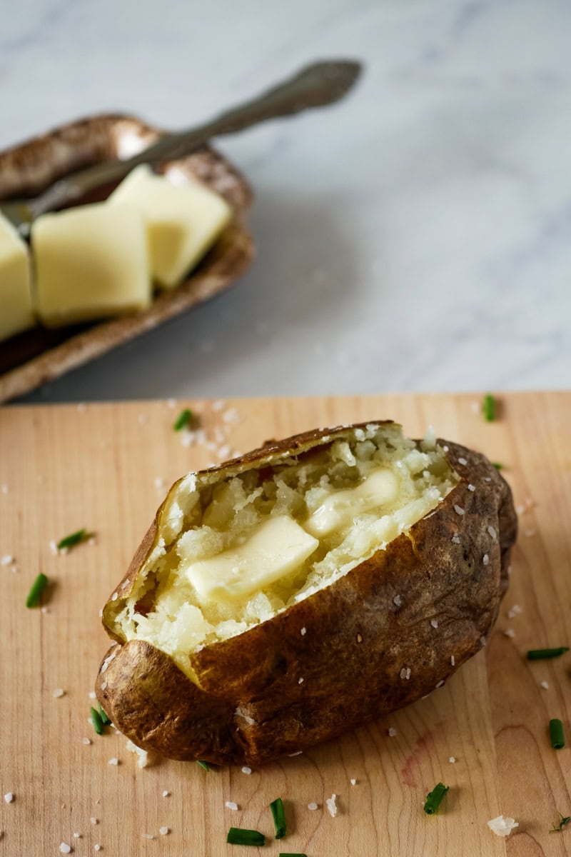 A baked potato split open with melting butter inside is on a wooden surface, garnished with coarse salt and chopped chives. Butter slices are in a dish in the background.