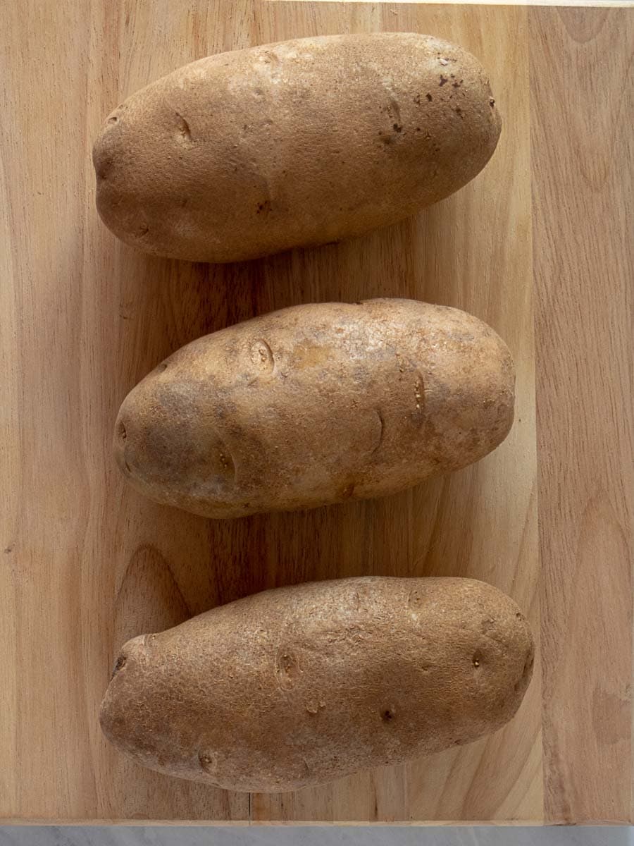 Three whole russet potatoes are arranged in a vertical line on a wooden cutting board.