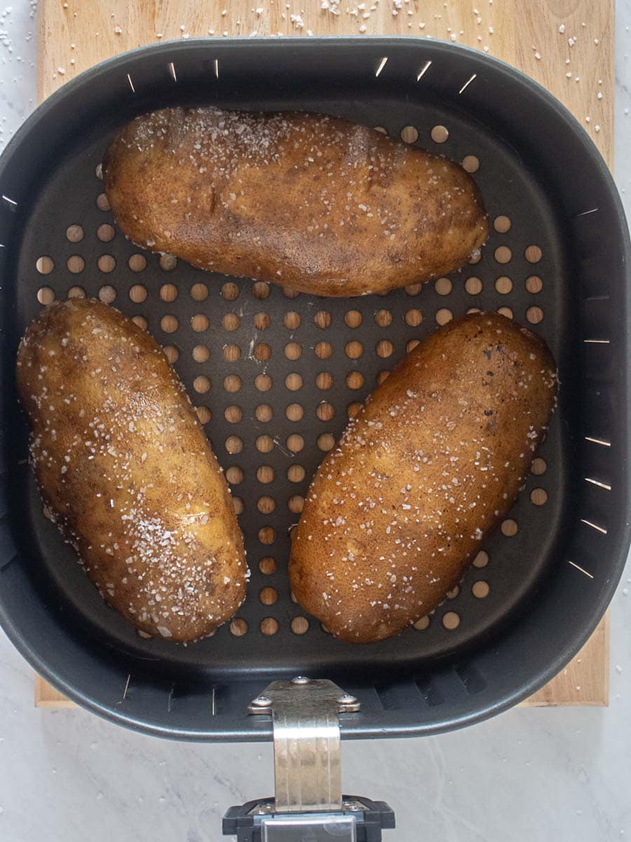 Three whole potatoes with salted skins are placed in an air fryer basket, ready to be cooked.