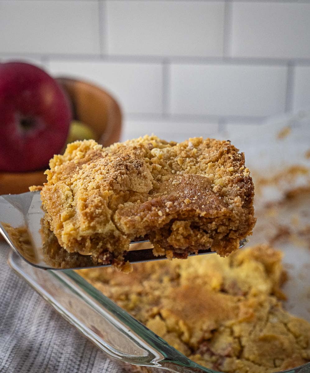 A metal spatula holds a square serving of crumbly apple dessert above a glass baking dish, with apples visible in the background.