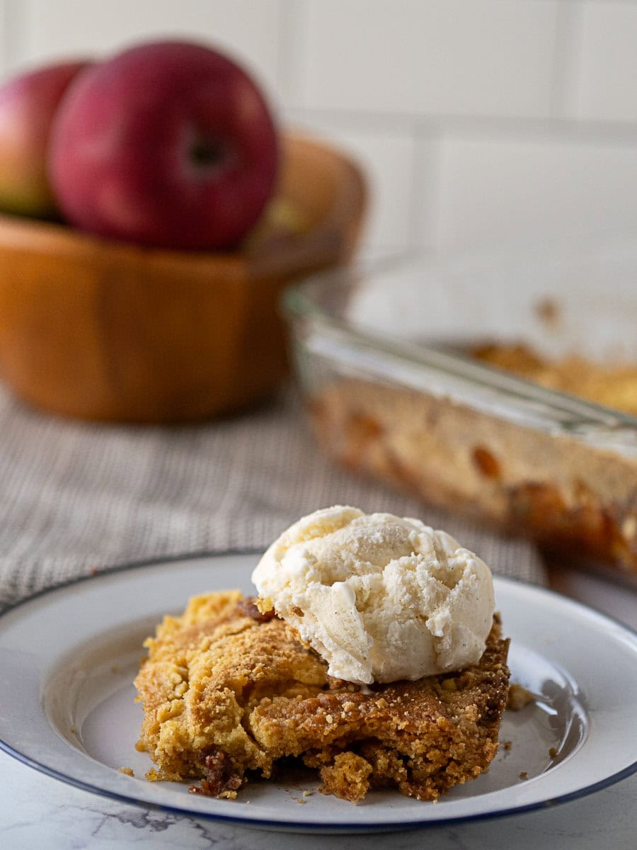 A serving of apple cobbler topped with a scoop of vanilla ice cream on a plate, with a baking dish and apples in the background.