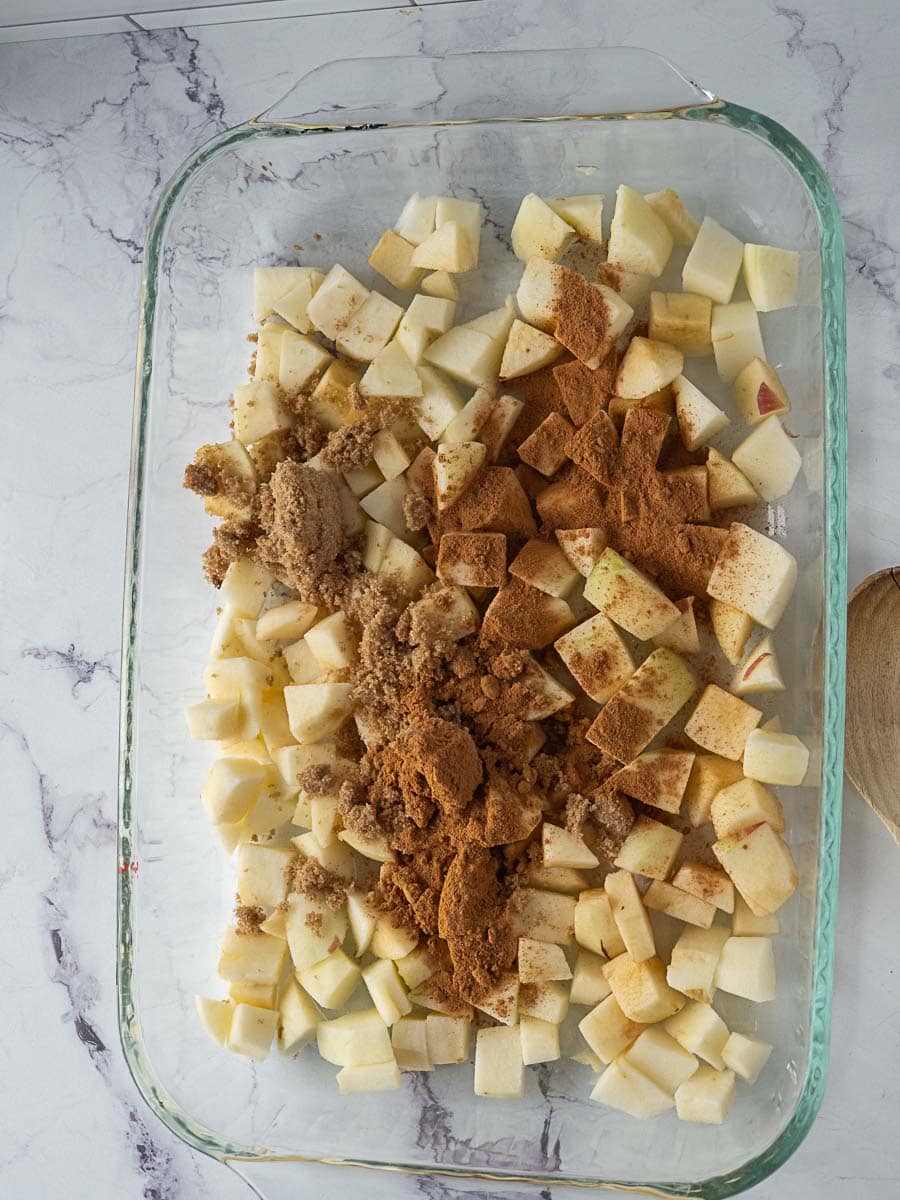 Glass baking dish with chopped apples, brown sugar, and cinnamon on a marble countertop, ready to be mixed.