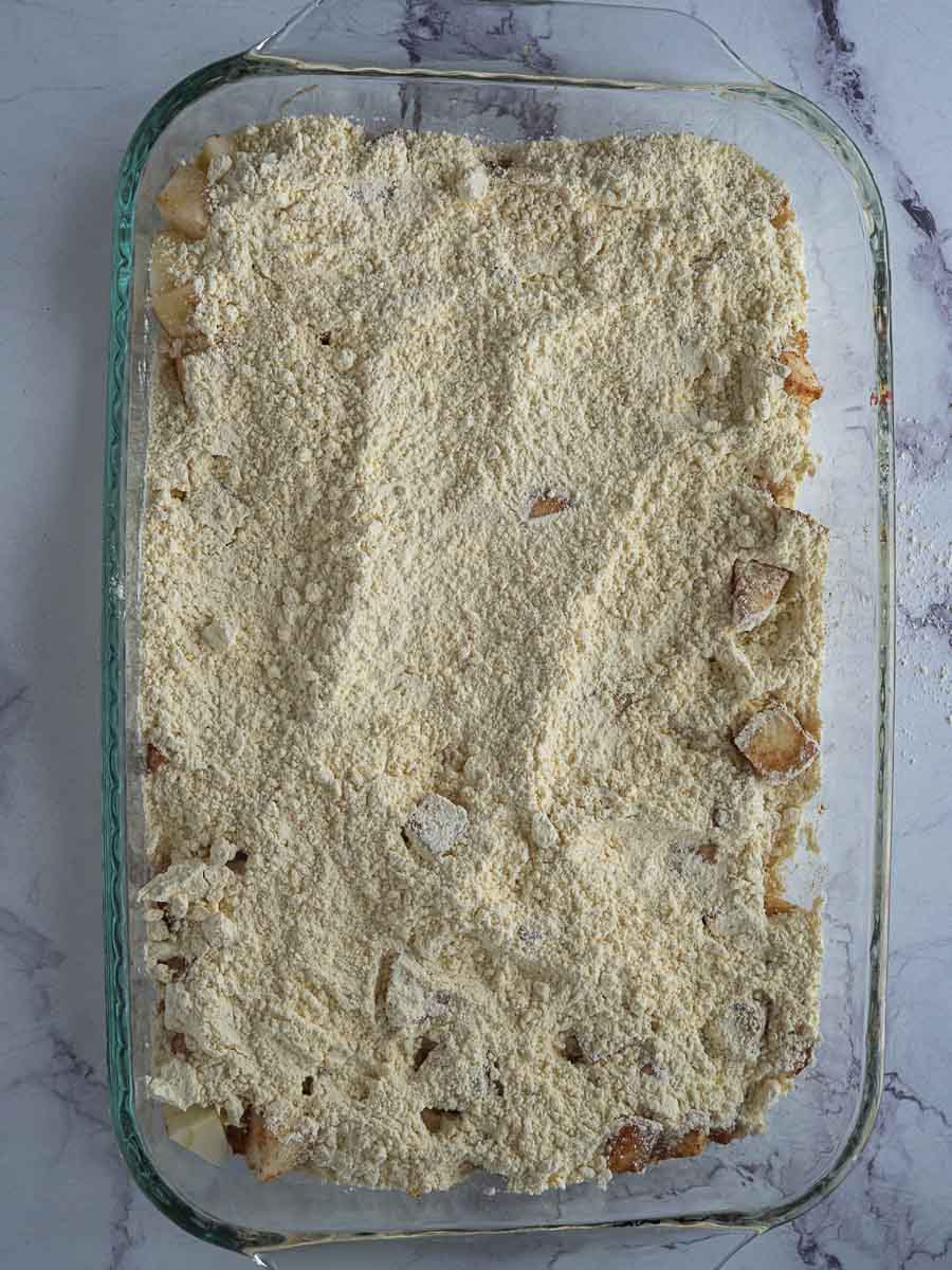 A glass baking dish filled with sliced apples and topped with an even layer of crumbly, uncooked streusel mixture, on a marble countertop.