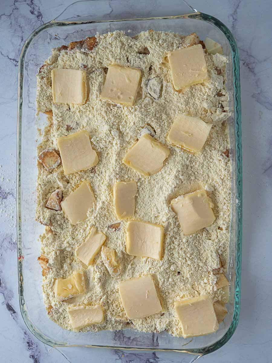 A glass baking dish filled with a crumbly mixture topped with several slices of butter, ready to be baked.