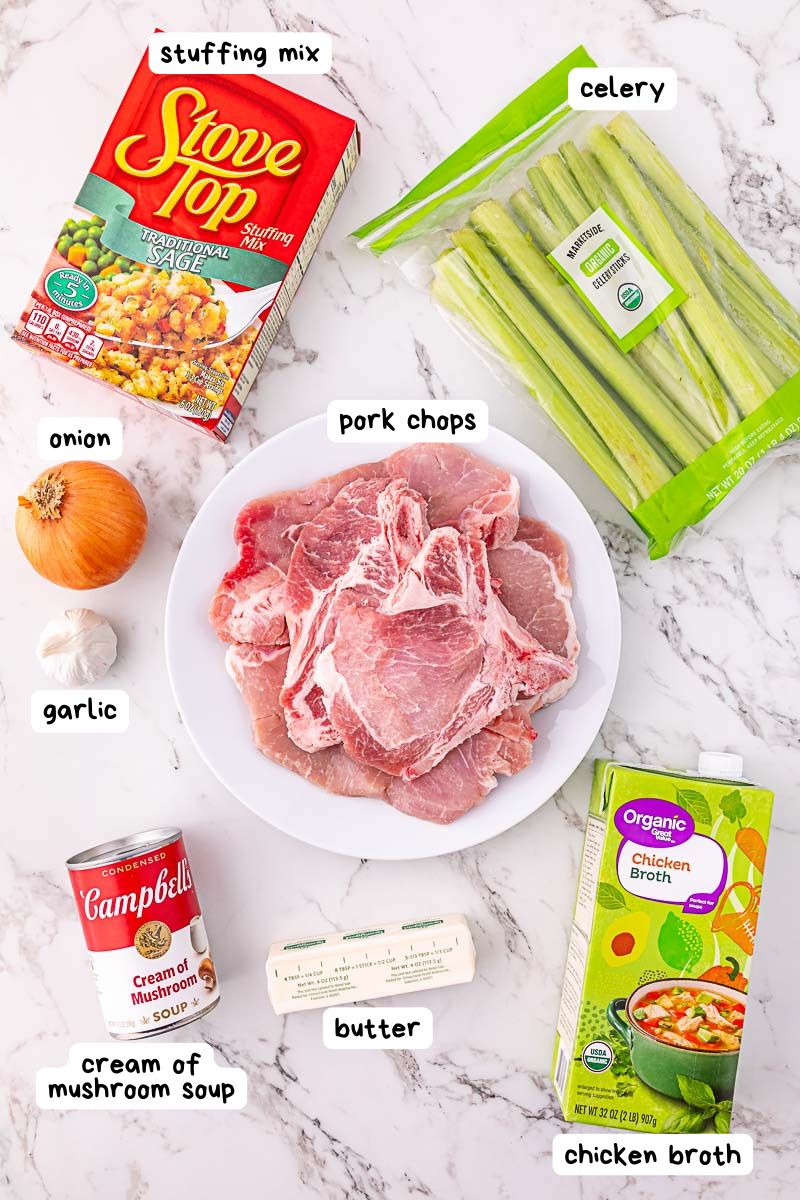 A plate of raw pork chops surrounded by stuffing mix, celery, onion, garlic, cream of mushroom soup, butter, and chicken broth on a white surface.