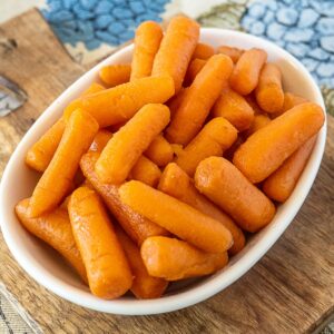 A white bowl filled with baby carrots sits on a wooden surface with a blue floral patterned cloth in the background.