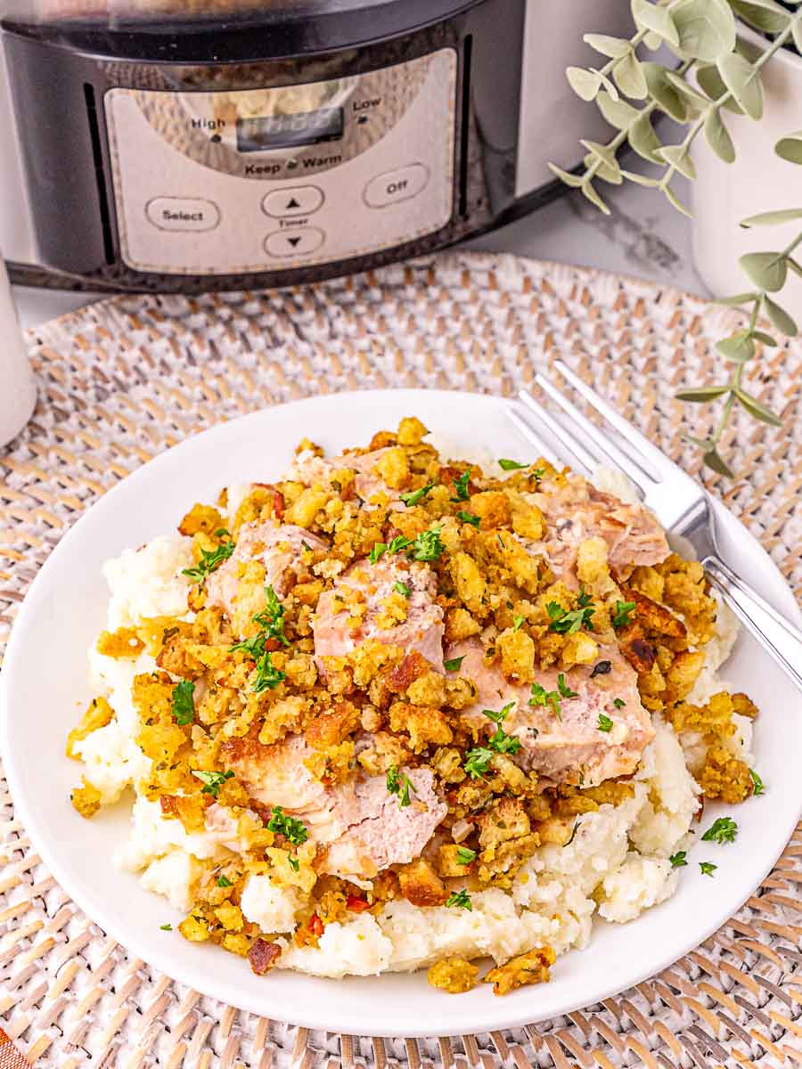 A plate of sliced turkey breast topped with stuffing and chopped herbs, served over mashed potatoes, with a fork and knife beside the plate and a slow cooker in the background.