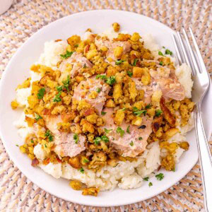 A plate of mashed potatoes topped with sliced chicken and seasoned stuffing, garnished with chopped parsley, with a fork and spoon on the side.