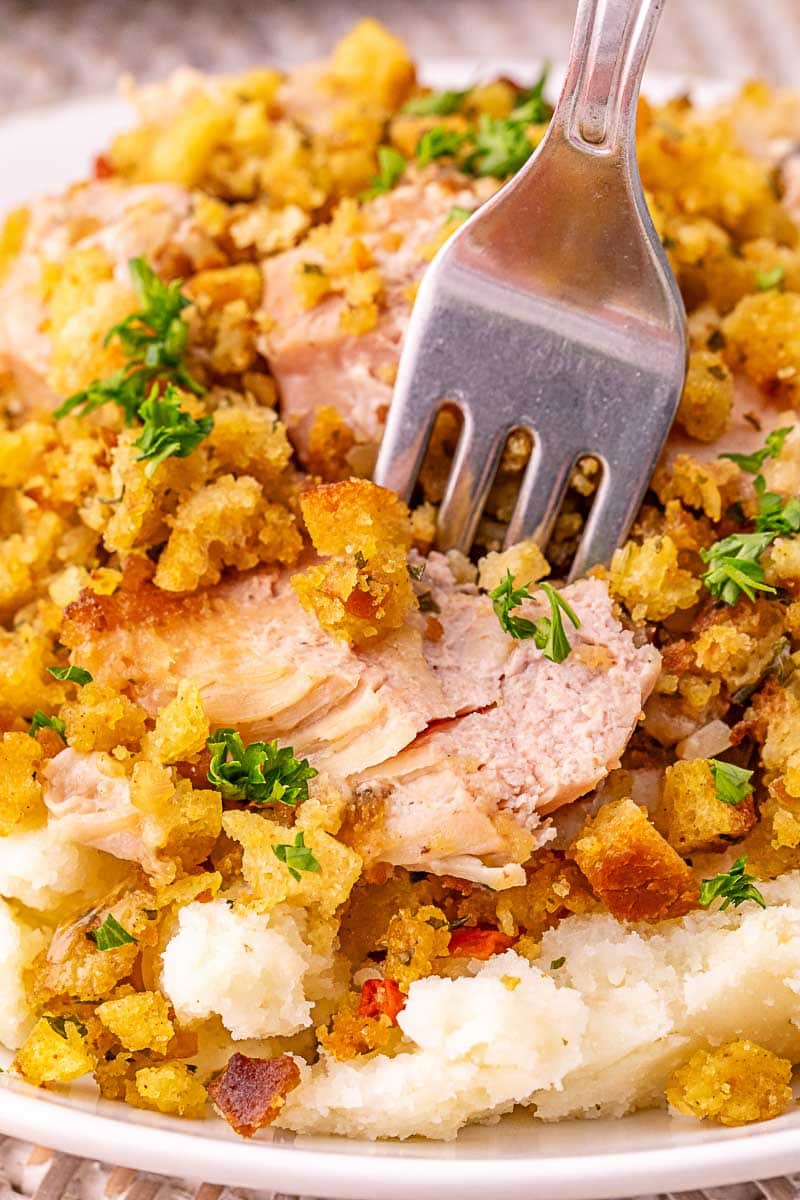 A fork pressing into a plate of mashed potatoes topped with shredded turkey, stuffing, and chopped parsley.