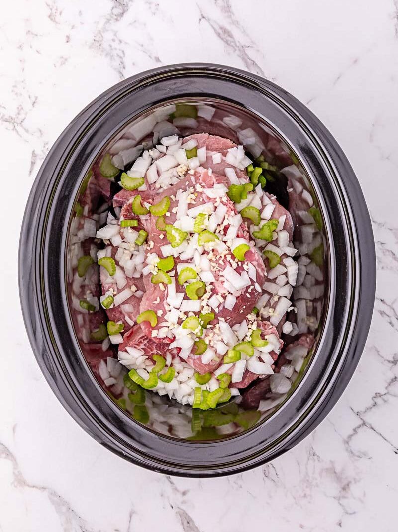 Raw beef slices, chopped onions, and green bell peppers are placed in a black slow cooker on a white marble surface.