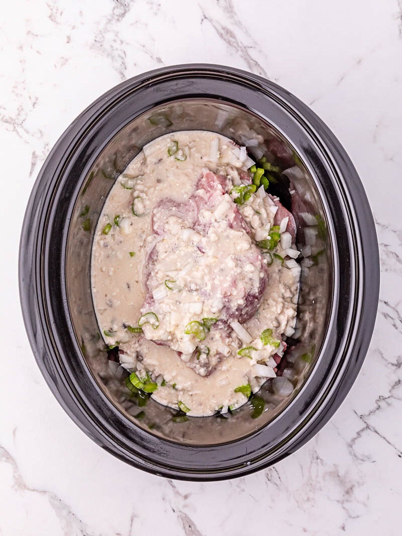 Raw meat, chopped onions, green peppers, and a creamy sauce placed in a black slow cooker on a white marble surface.