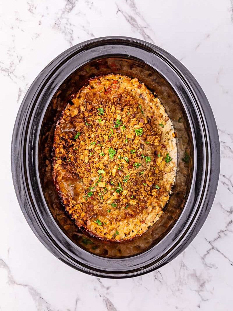 Overhead view of a cooked casserole topped with breadcrumbs and herbs inside a black slow cooker on a marble surface.