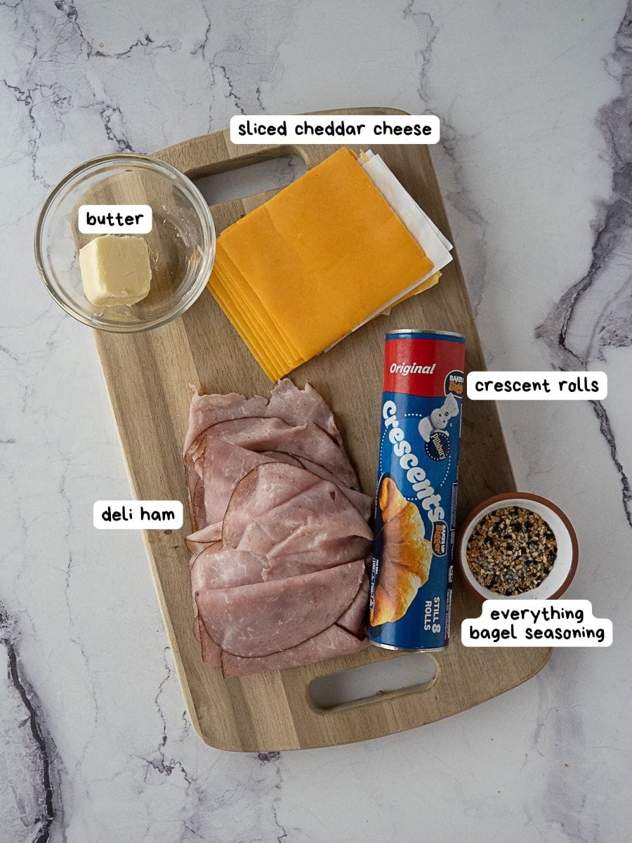 A wooden board with butter in a glass bowl, sliced cheddar cheese, a can of crescent rolls, deli ham slices, and a small bowl of everything bagel seasoning.