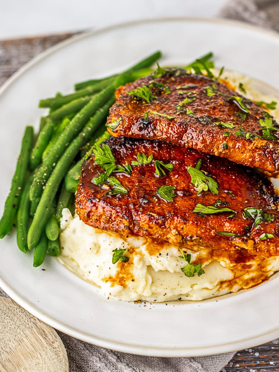 Two seasoned pork chops served on mashed potatoes with green beans on the side, garnished with chopped parsley.