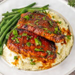 Two pieces of seasoned, grilled chicken breast on mashed potatoes, garnished with parsley, served with green beans on a white plate.