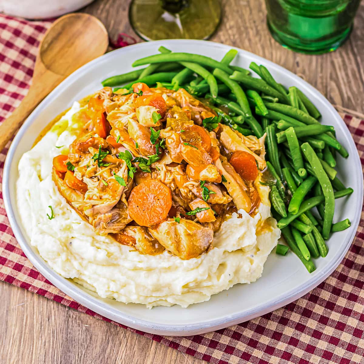 A plate of mashed potatoes topped with shredded chicken and carrots in sauce, served with steamed green beans on the side.