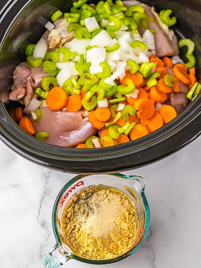 Uncooked chicken, chopped carrots, celery, and onions in a slow cooker; a measuring cup with powdered seasonings and broth on the side.