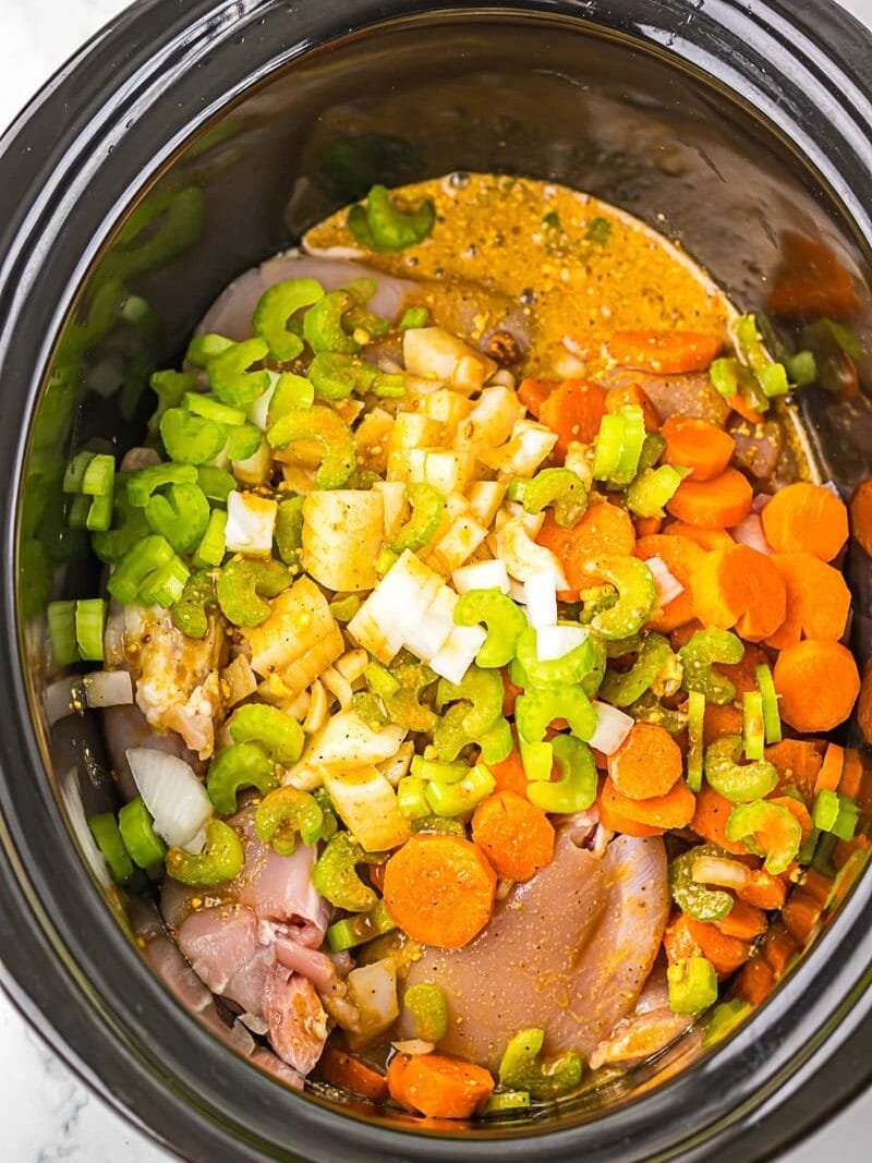 Raw chicken, chopped carrots, celery, and onions with seasoning and broth in a slow cooker, ready for cooking.