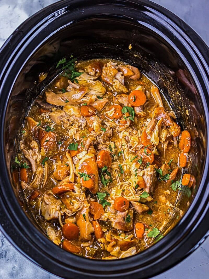 A slow cooker filled with shredded meat, sliced carrots, and a savory sauce, garnished with chopped herbs.