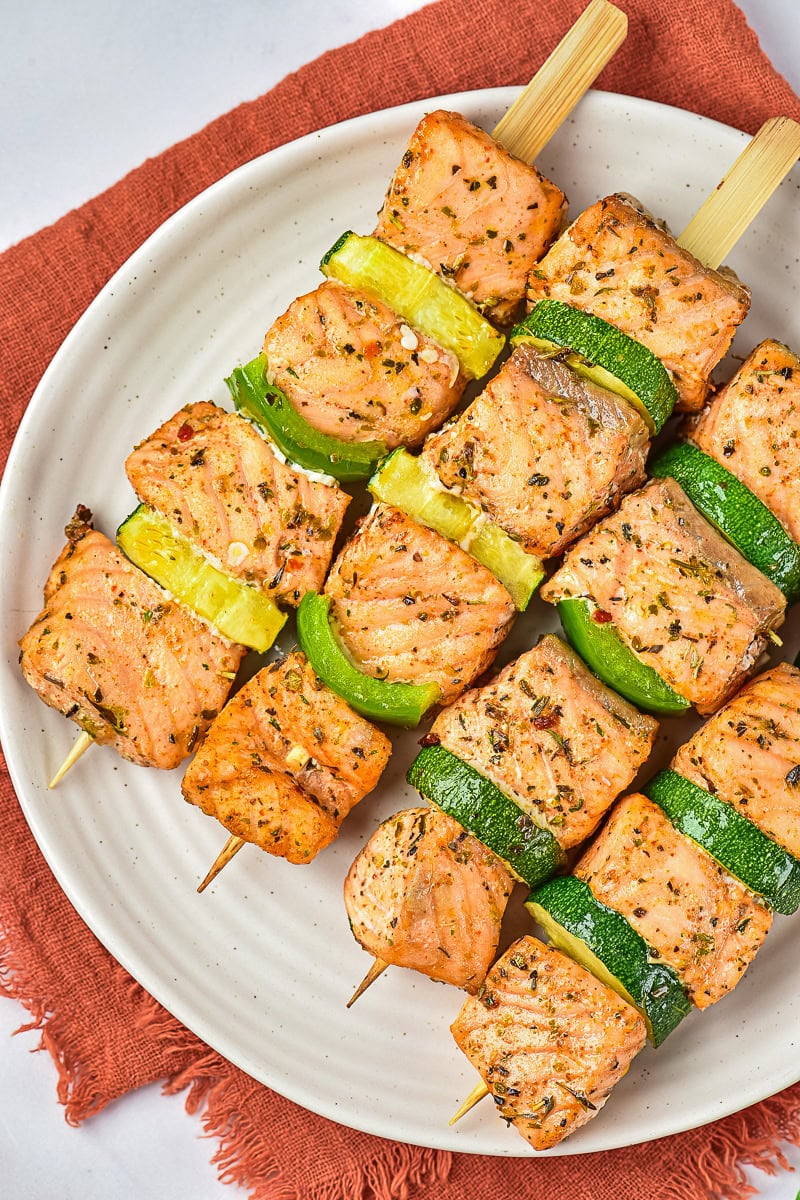 Three skewers with seasoned salmon cubes, green bell pepper, and zucchini are arranged on a white plate atop a rust-colored napkin.