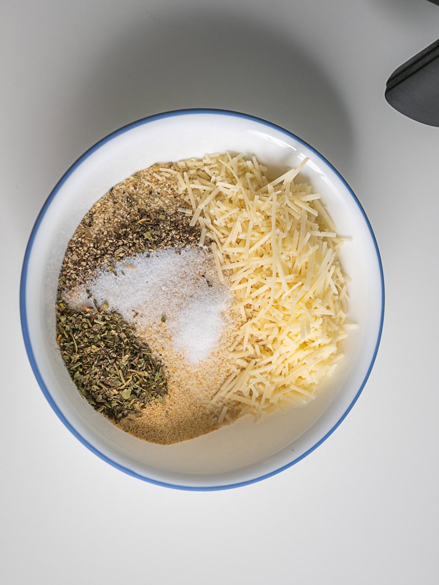A white bowl containing shredded cheese, dried herbs, black pepper, salt, and breadcrumbs&mdash;perfect toppings for air fryer chicken parmesan&mdash;viewed from above on a white surface.