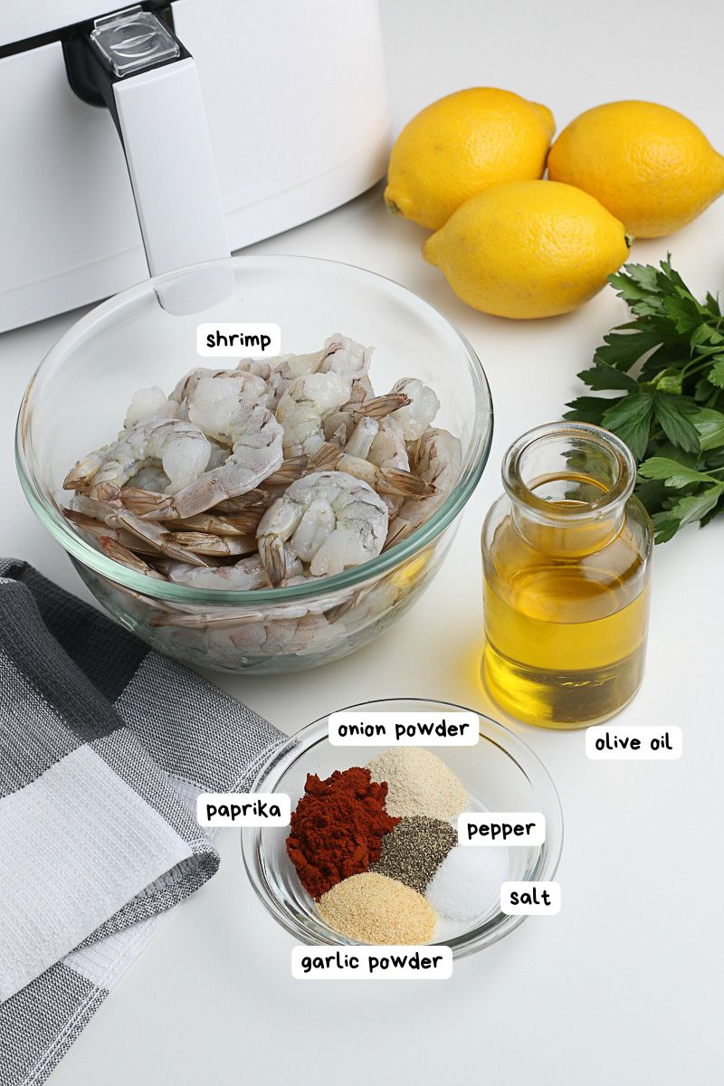 Bowl of raw shrimp, olive oil, spices (paprika, onion powder, garlic powder, salt, pepper), lemons, and parsley on a countertop next to an air fryer.