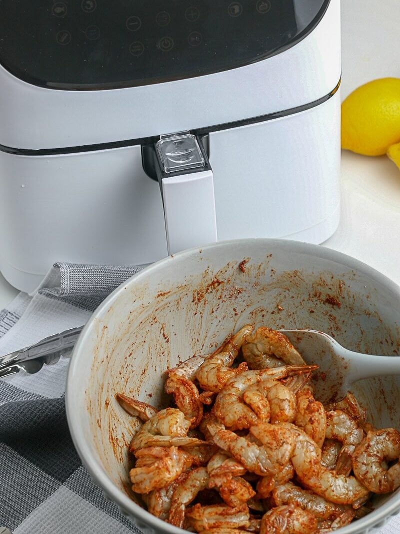 A bowl of seasoned raw shrimp with a spoon in front of a white air fryer, with lemons and a kitchen towel nearby.