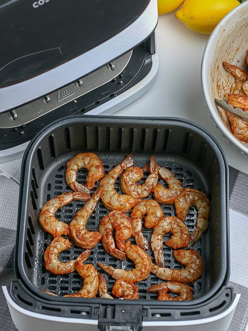 Raw seasoned shrimp arranged in a single layer in the basket of an open air fryer, with a bowl of shrimp and lemons nearby on the counter.