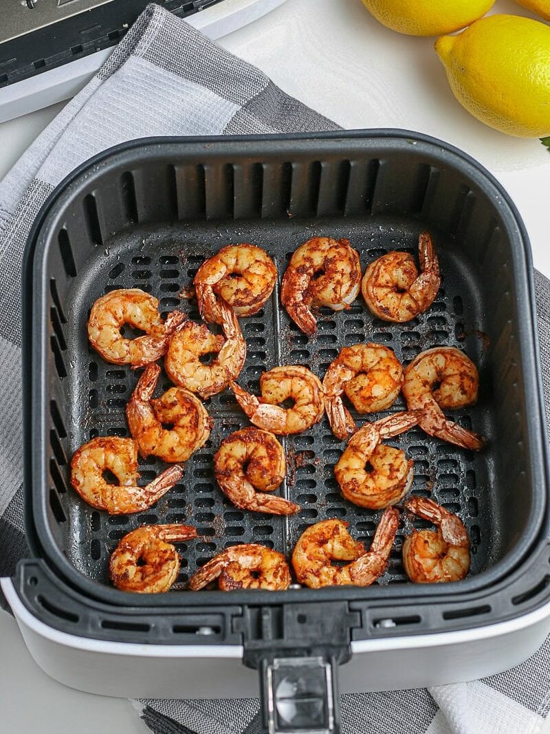 Cooked shrimp arranged in an air fryer basket, with two whole lemons and a striped cloth nearby on a white surface.