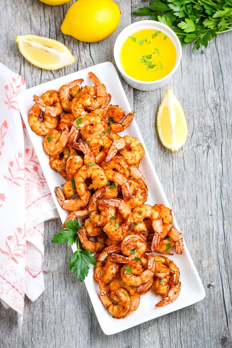A rectangular plate of cooked shrimp garnished with parsley, served with lemon wedges and a cup of yellow dipping sauce on a wooden table.