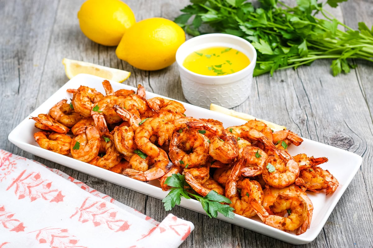 Grilled shrimp piled on a rectangular white plate, garnished with parsley, with lemon wedges, fresh parsley, two whole lemons, and a small bowl of melted butter on a wooden table.