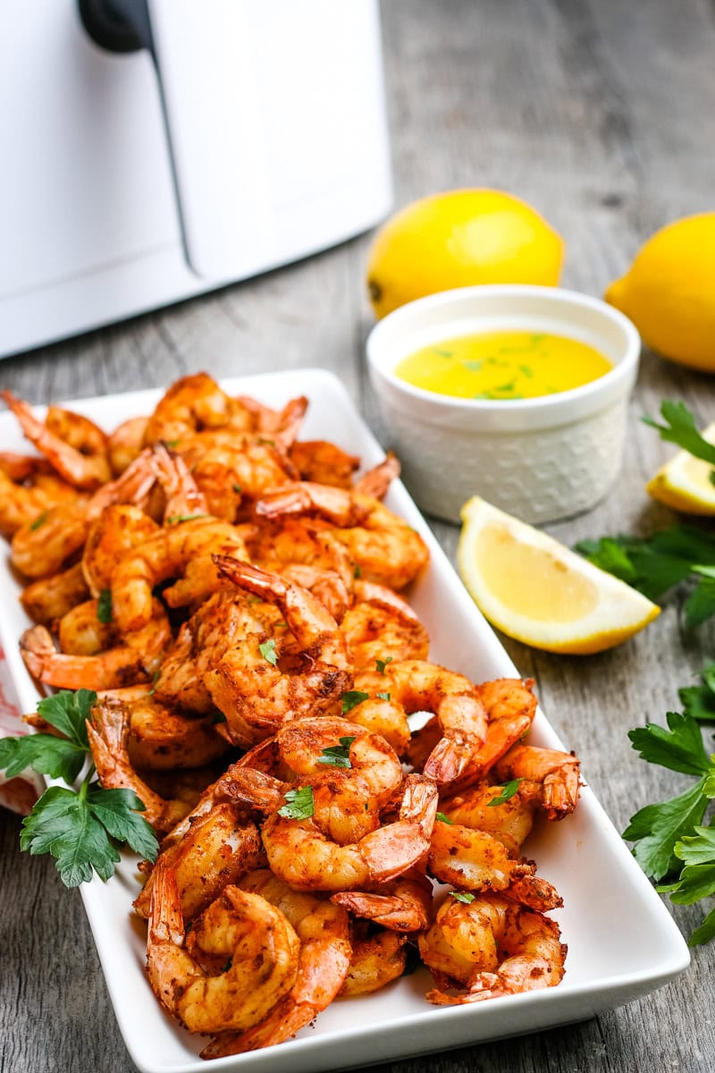 A rectangular white plate filled with seasoned cooked shrimp, garnished with parsley, next to lemon wedges, fresh parsley, a cup of yellow sauce, and two whole lemons.