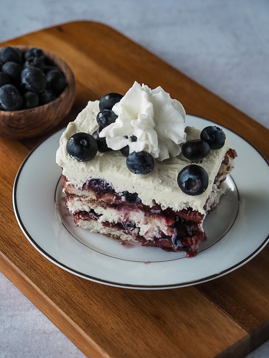 A slice of blueberry dessert with whipped cream and blueberries on top, served on a white plate with a bowl of blueberries nearby.