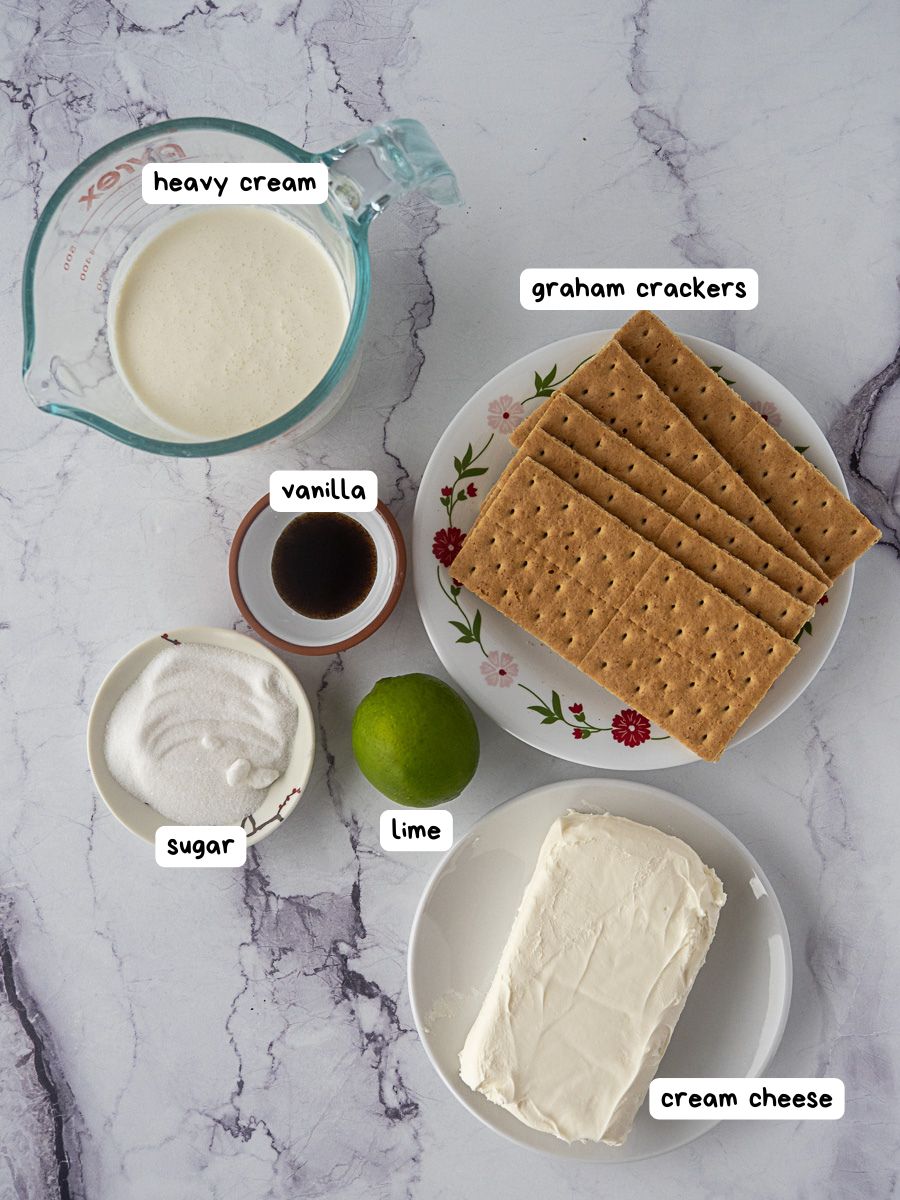 Top-down view of ingredients for a dessert: heavy cream, graham crackers, cream cheese, sugar, vanilla extract, and a lime arranged on a marble surface.