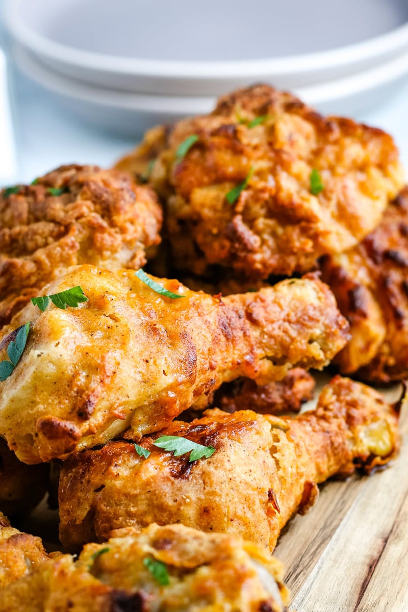 A close-up of crispy baked chicken drumsticks garnished with chopped herbs, stacked on a wooden surface with plates in the background.