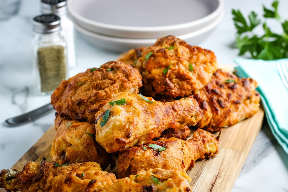 A pile of golden, crispy fried chicken pieces garnished with parsley on a wooden board, with plates, napkins, and seasoning shakers in the background.