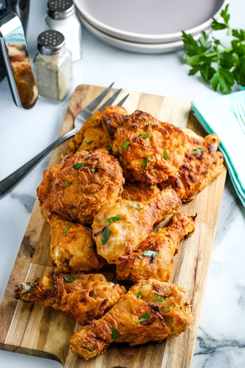 A wooden board topped with pieces of golden fried chicken, garnished with herbs. Plates, utensils, salt, pepper shakers, and fresh parsley are in the background.