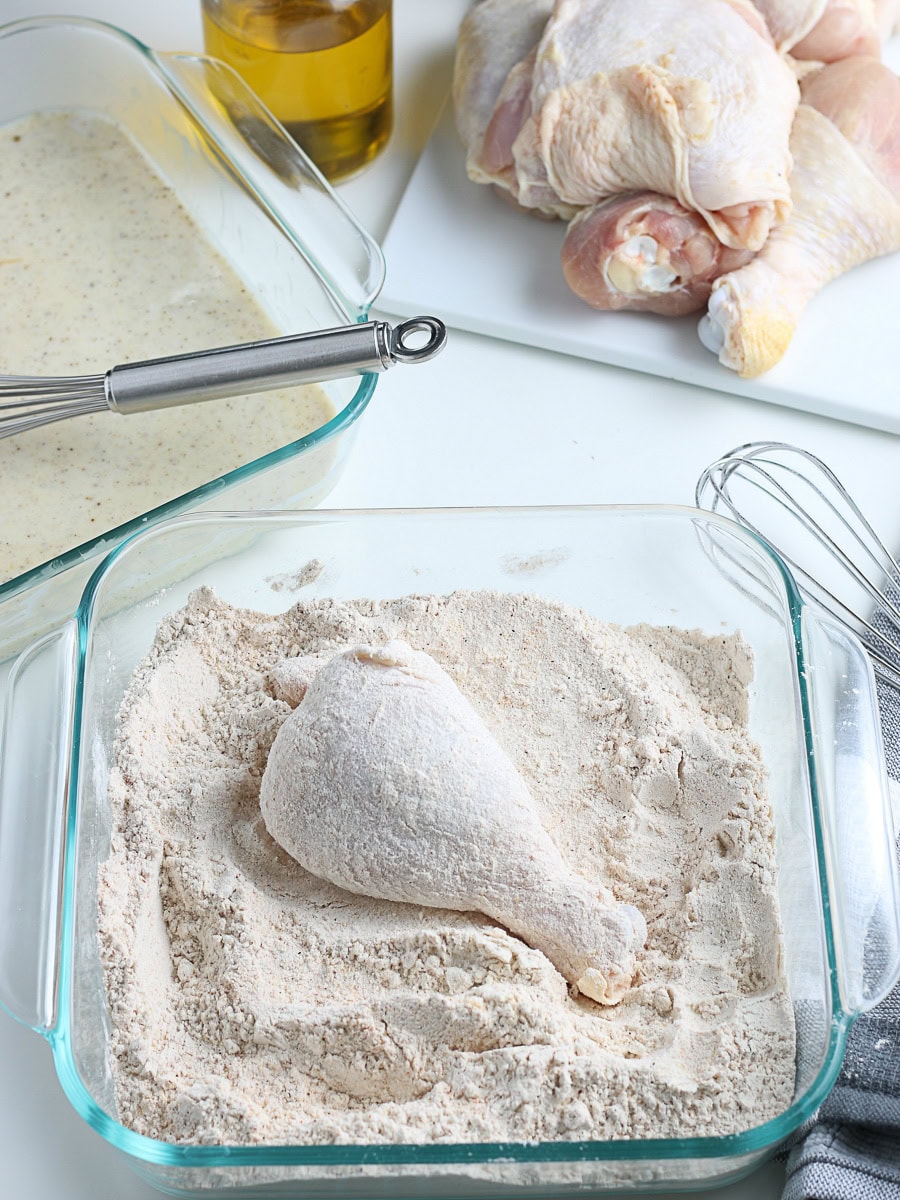 A raw chicken drumstick coated in flour sits in a glass dish, with a bowl of batter, raw chicken pieces, a whisk, and a bottle of oil nearby.