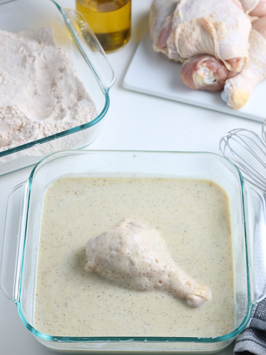 A raw chicken drumstick is being dipped in a seasoned batter, with flour mixture, oil, and more raw chicken pieces visible nearby.